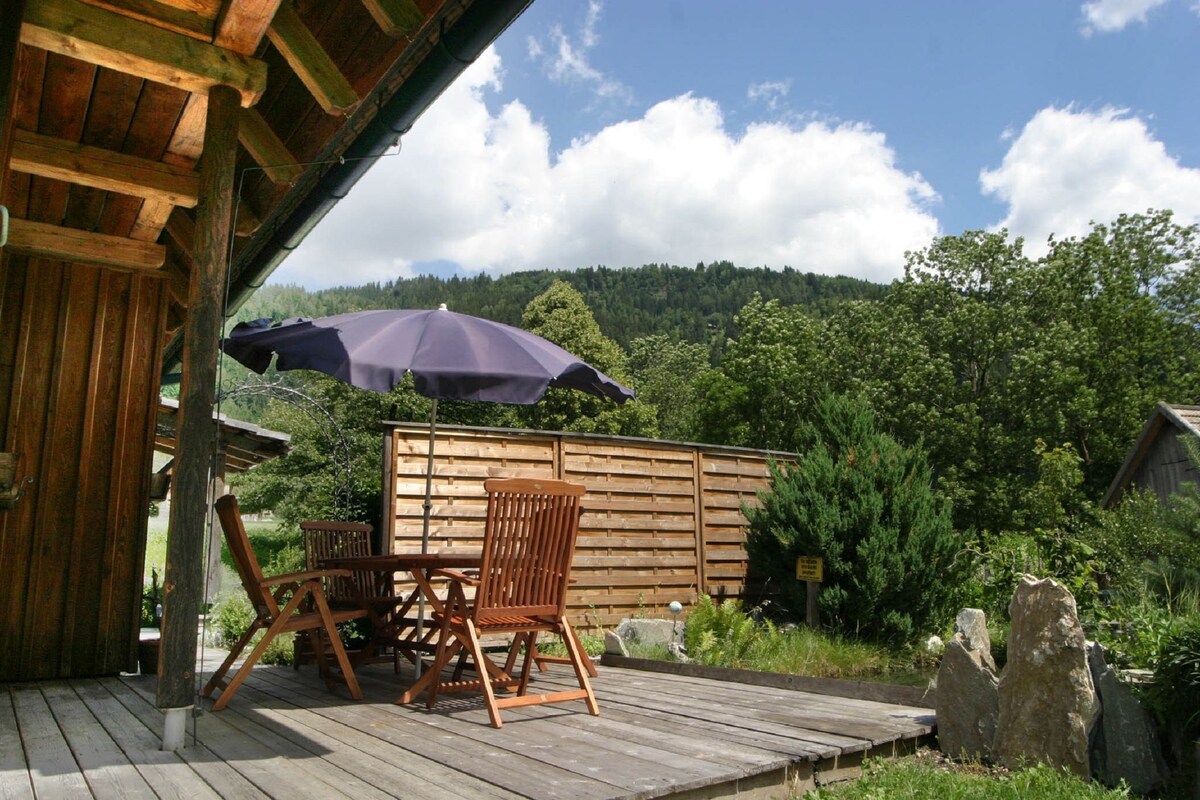 A sunny terrace is depicted with wooden furniture, including two chairs and a small table. A large umbrella provides shade, while a wooden fence and lush greenery frame the space, creating a relaxed outdoor environment with views of the surrounding mountains.