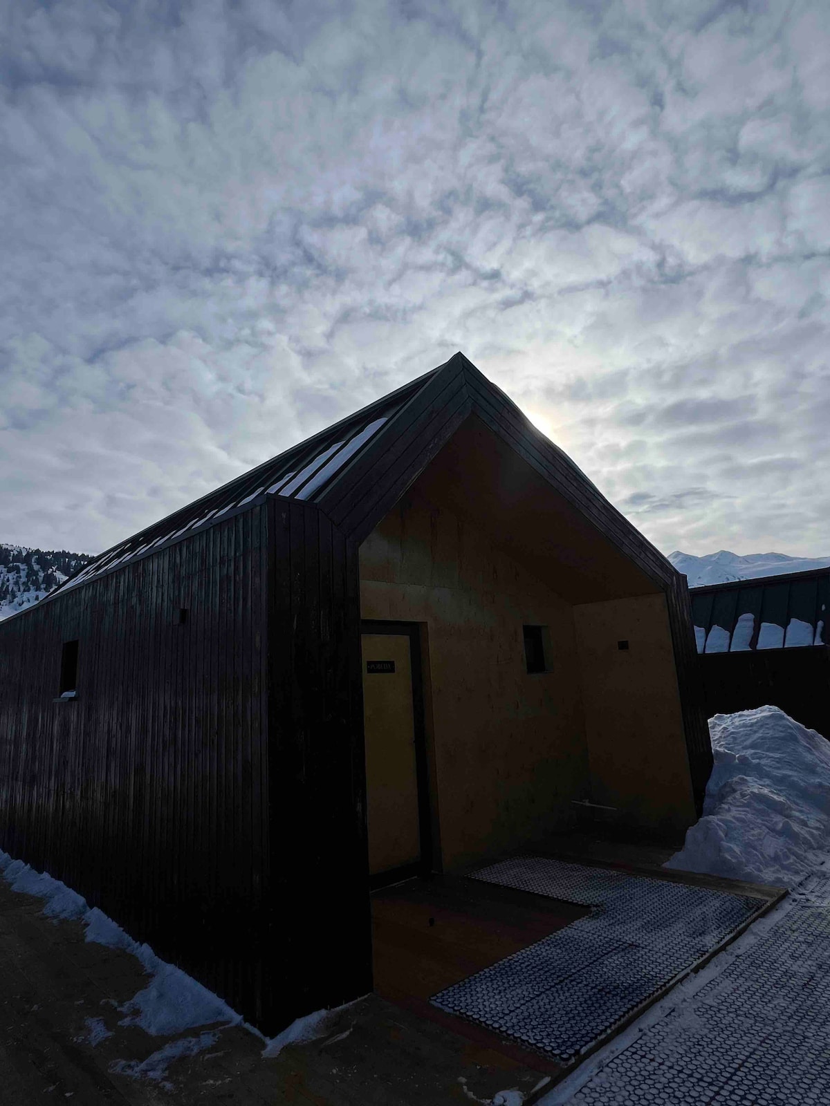 The exterior of a modern cottage is captured under a cloudy sky, showcasing a unique triangular roof. The entrance is highlighted with a welcome mat, and snow subtly frames the base, creating a serene winter ambiance.