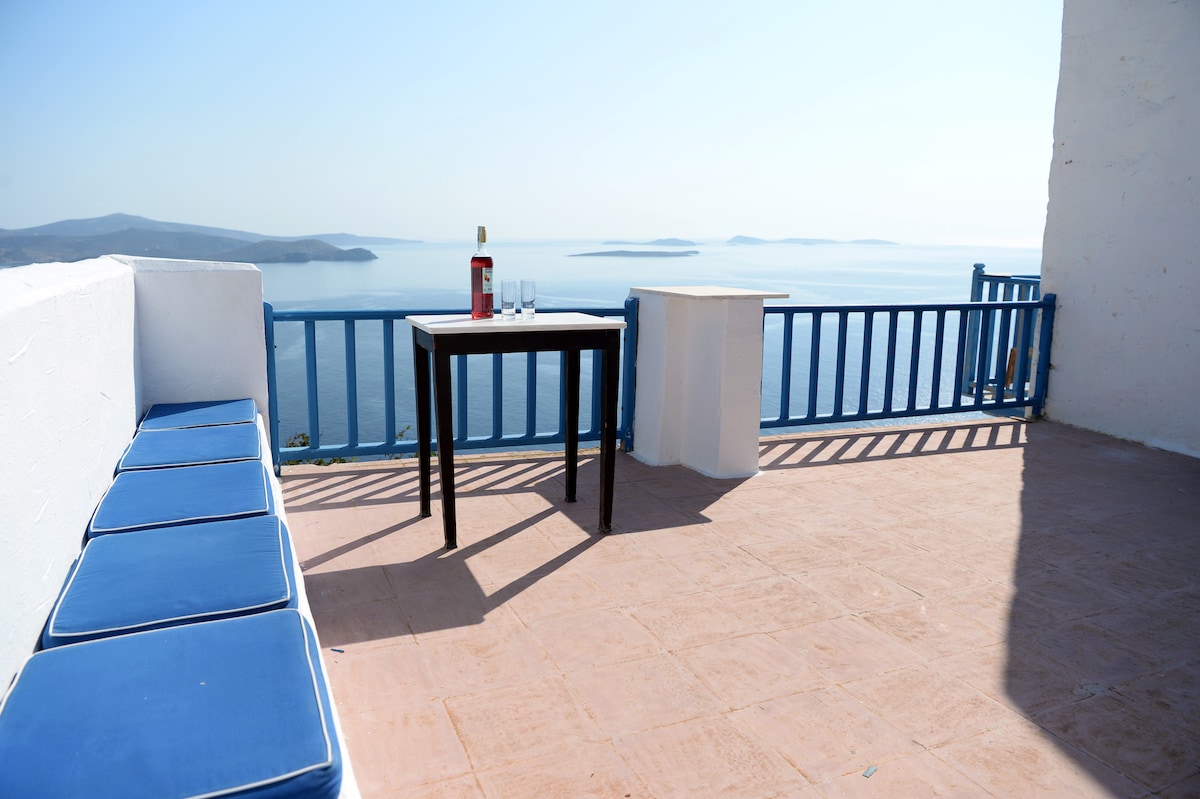 A spacious terrace offers stunning views of the Aegean Sea, featuring a table set for two with wine glasses and a bottle. Blue cushioned seating lines one side, inviting relaxation while enjoying the scenic backdrop of distant islands.