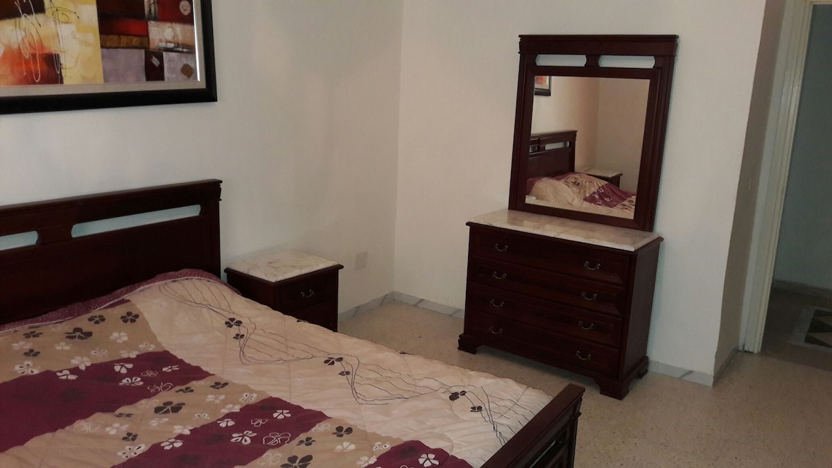 A spacious bedroom is presented with a wooden bed frame featuring a patterned quilt. A dark wood dresser and nightstand are placed near the bed, with a matching mirror above the dresser. Soft lighting enhances the calm environment.