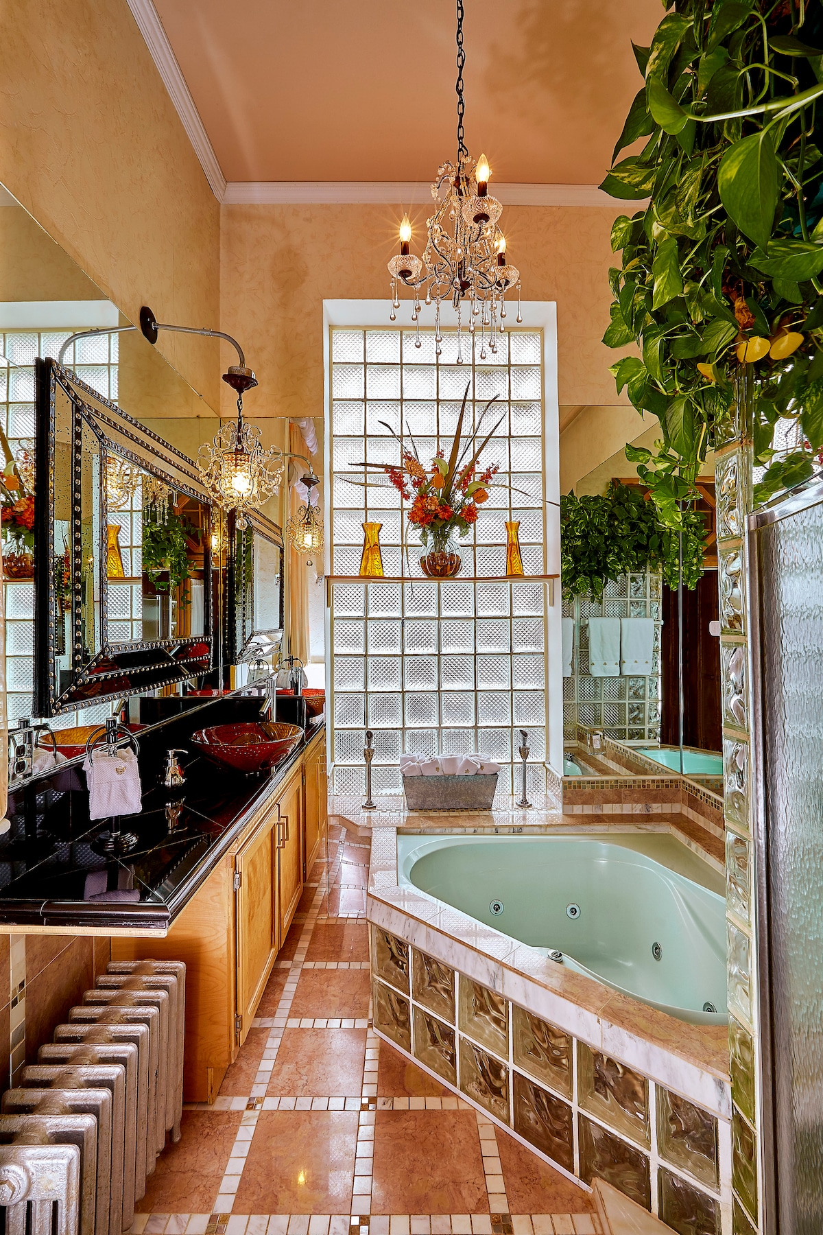 A spacious bathroom features a heart-shaped Jacuzzi tub beside marble-tiled flooring. Elegant lighting is provided by a chandelier, while large mirrors enhance the room's brightness. Decorative plants and vases filled with flowers add a touch of color against the glass block wall.