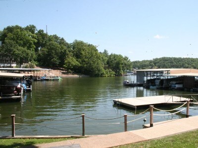 A serene view of the cove is presented, featuring calm waters bordered by lush greenery. Several boat docks are visible along the shoreline, with boats gently moored. The clear sky and distant hills create a peaceful backdrop to this inviting lakeside setting.