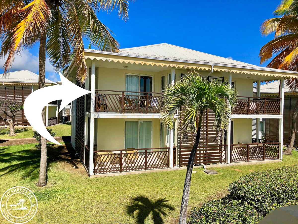 A two-story bungalow is surrounded by palm trees and landscaped greenery. The balcony features wooden railings and overlooks the lawn area, offering a shaded spot to enjoy the tropical environment. Large windows allow natural light into the living spaces.