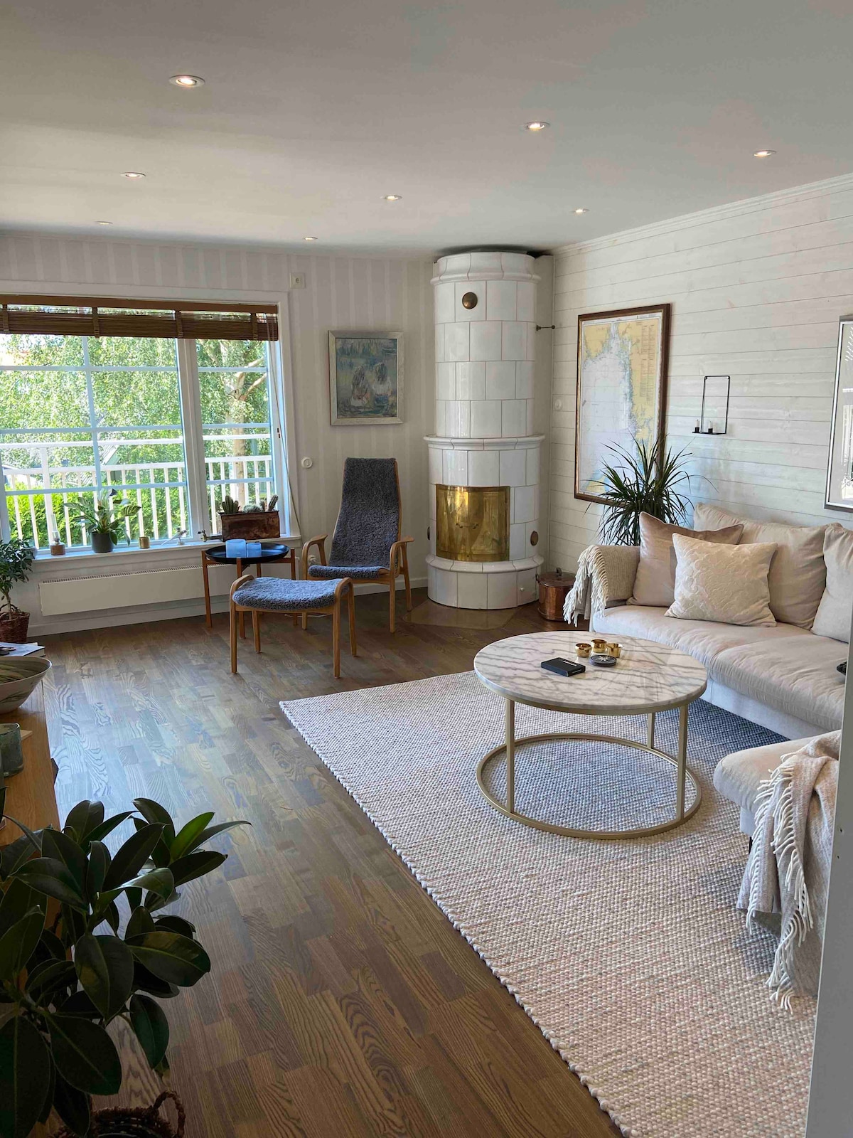 A bright living area features a comfortable sofa and a round coffee table, with a cozy chair positioned nearby. Large windows provide natural light and views of the outdoors. A decorative stone fireplace stands in the corner, complemented by indoor plants.