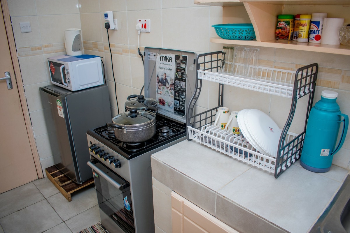 A modern kitchen setup features a compact stove with a pot, complemented by a microwave and a small refrigerator. A drying rack houses plates and cups, while a blue water container adds a touch of color. Neutral-toned cabinets and countertops support the functional design.