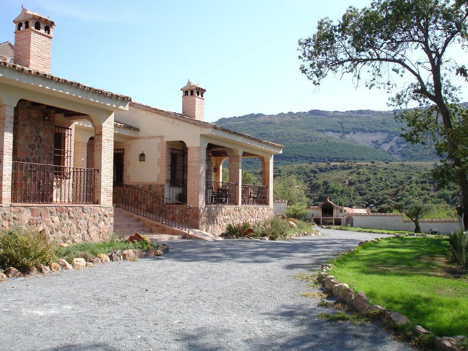 The exterior of a spacious villa is depicted, featuring a gravel driveway lined with stones. The structure showcases a combination of stone and plaster walls, with balconies and two brick chimneys. A lush green lawn and mountainous landscape provide a serene backdrop.