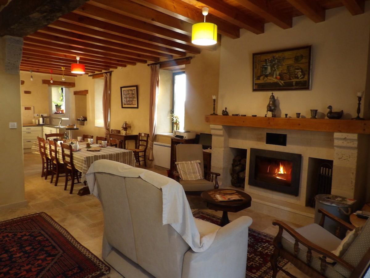 A welcoming living space features a fireplace with logs stacked beside it. Comfortable seating is arranged around a central coffee table. Warm lighting is provided by overhead lamps, enhancing the rustic wooden beams and neutral walls throughout the room.