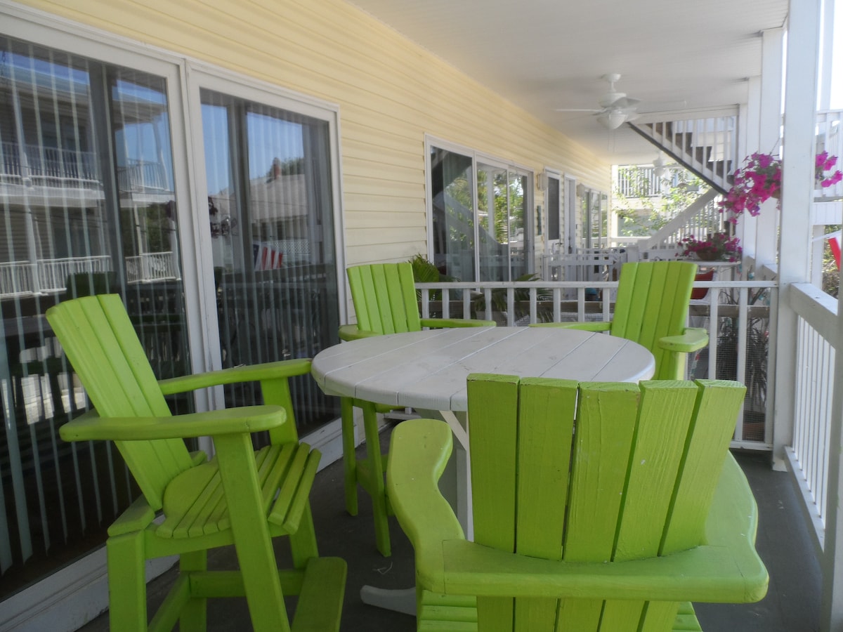 A spacious outdoor deck is furnished with a circular white table surrounded by four bright green adirondack chairs. The area is bordered by sliding glass doors that provide access to the interior, with flowering plants adding a touch of color.
