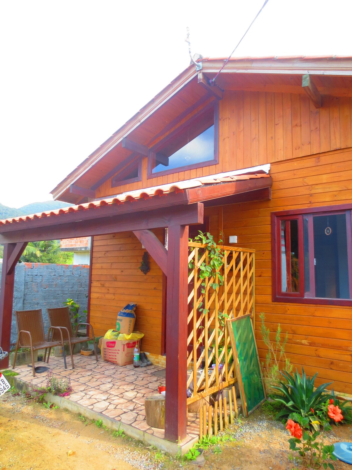 The image shows the exterior of a wooden house featuring a covered patio area with two chairs. A decorative trellis is visible, accompanied by greenery and a pathway leading to the entrance. Large windows allow natural light into the space.