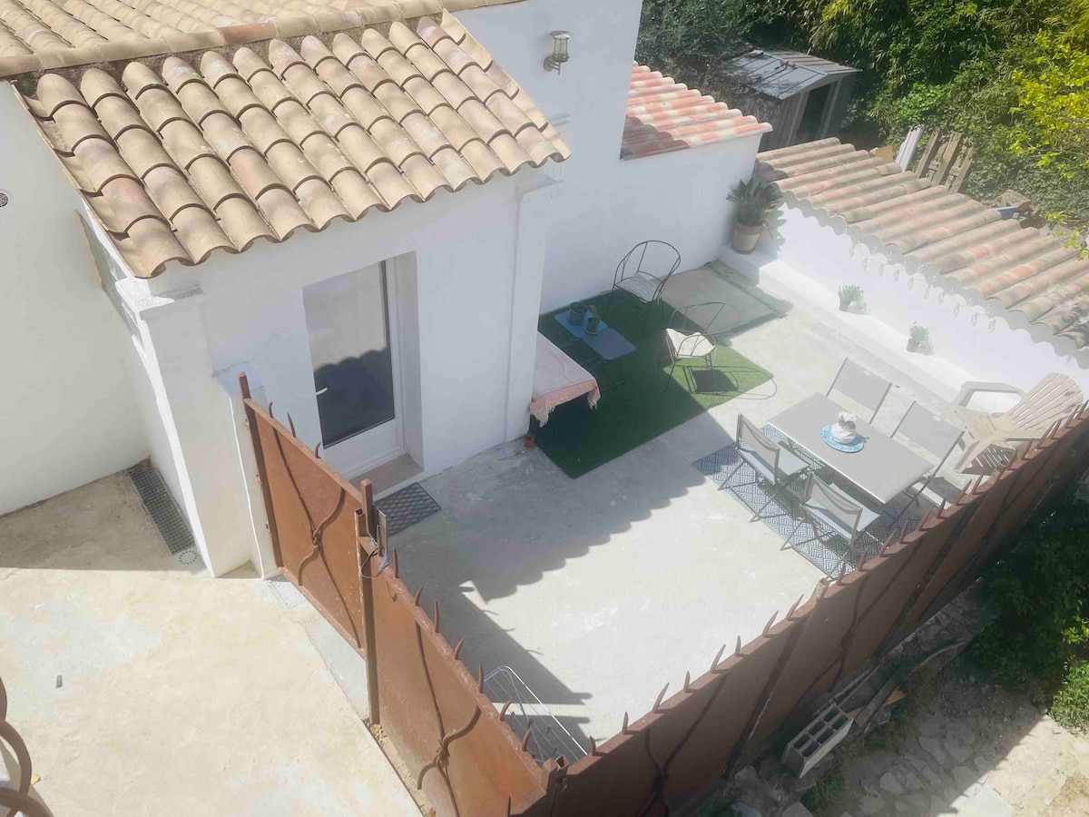 An outdoor terrace features a small table set with chairs, surrounded by a green area and potted plants. The restful seating arrangement is complemented by a view of the nearby walls and vegetation. Sunlight illuminates the space, and a tiled roof is visible above.