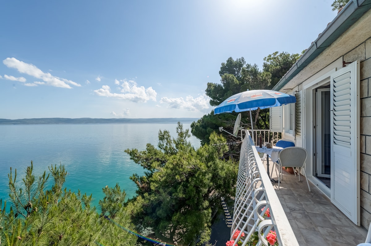 A spacious balcony offers a panoramic view of the tranquil sea, surrounded by lush greenery. Sunlight reflects off the water, providing a serene atmosphere. A blue parasol shades a small table and two chairs, inviting guests to relax and enjoy the picturesque setting.