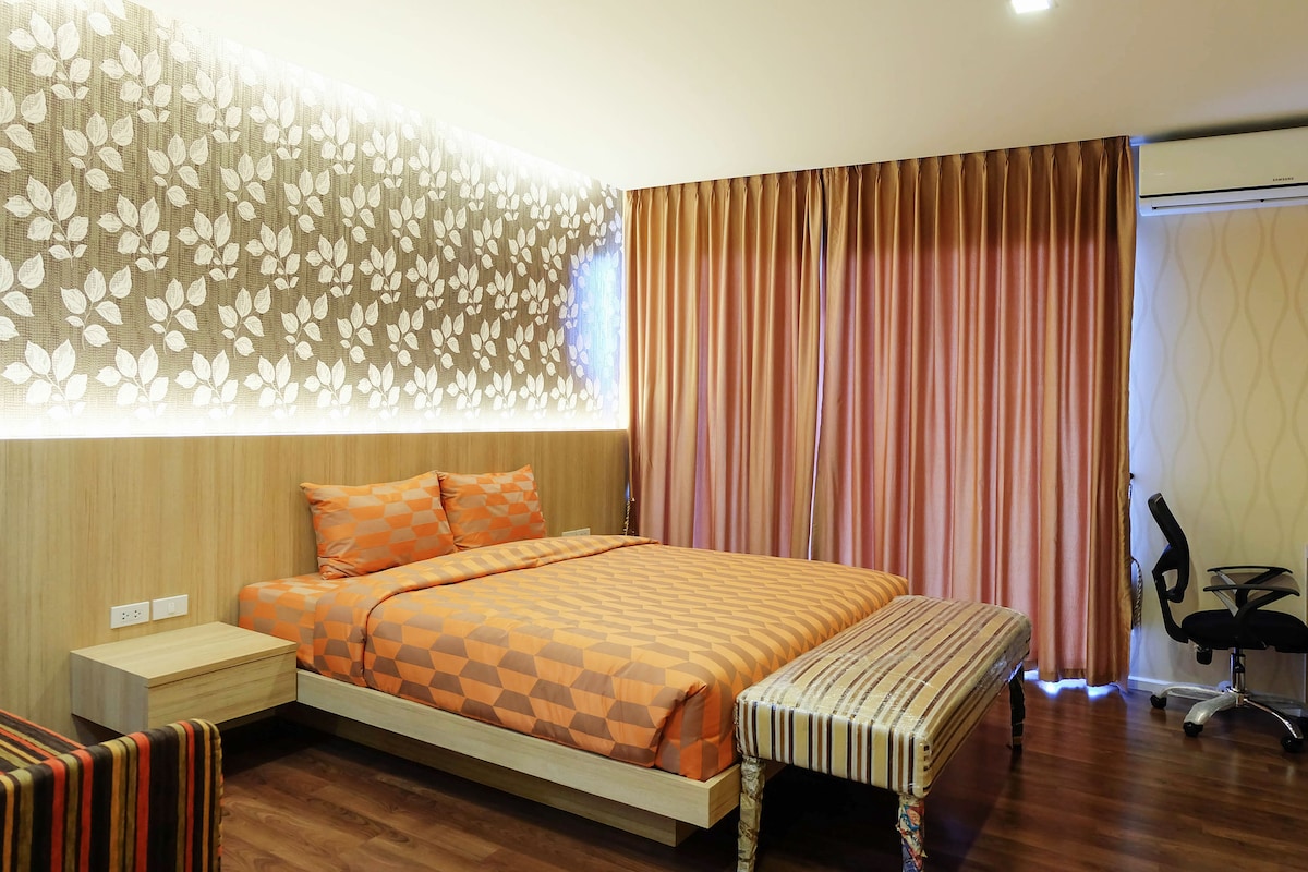 A well-lit bedroom features a spacious bed with an orange geometric patterned duvet. The wall behind is adorned with leaf motifs, while large curtains in warm tones complement the decor. A striped bench sits at the foot of the bed, and a modern chair is positioned nearby.