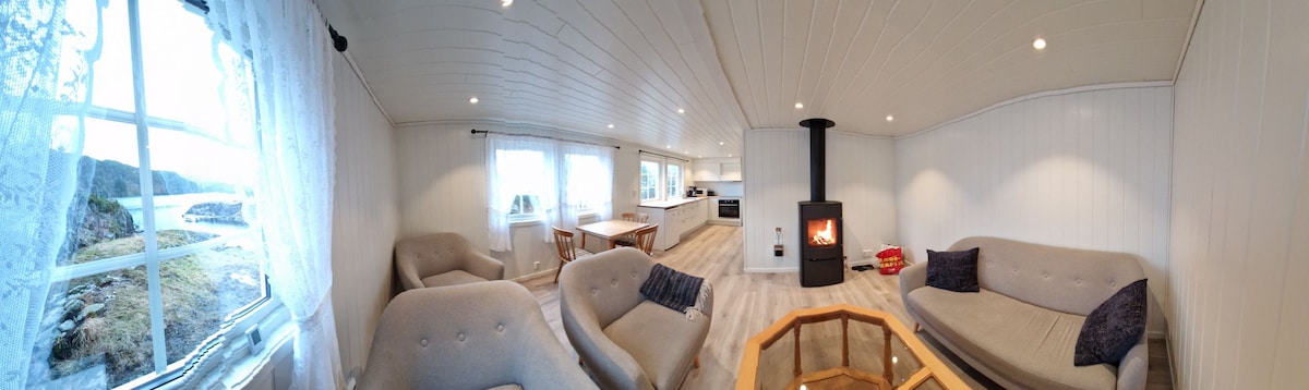 An open-plan living space showcases a wood-burning stove as a central feature. Natural light floods in through large windows, enhancing the light wood flooring. Soft seating arrangements are complemented by decorative pillows, while a dining area is visible in the background.