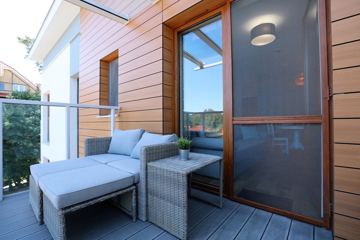 An outdoor seating area features a comfortable gray chaise lounge and a small table, set on a wooden deck. A potted plant adds a touch of greenery, while large sliding doors provide access to the interior space and views of the surrounding nature.