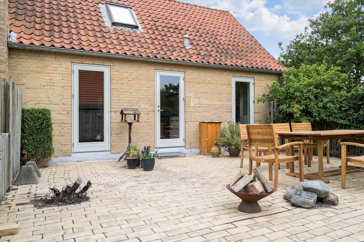 A spacious outdoor patio showcases a brick surface surrounded by greenery. A wooden dining table with chairs is positioned nearby, complemented by potted plants and a small grill. The brick exterior of the house adds a rustic charm, with windows providing natural light.