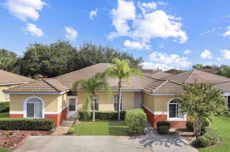 A single-story home with a tiled roof is surrounded by well-maintained landscaping, featuring lush green lawns and palm trees. Large windows enhance the exterior's bright yellow and muted red colors, while the inviting front path provides access to the entrance.