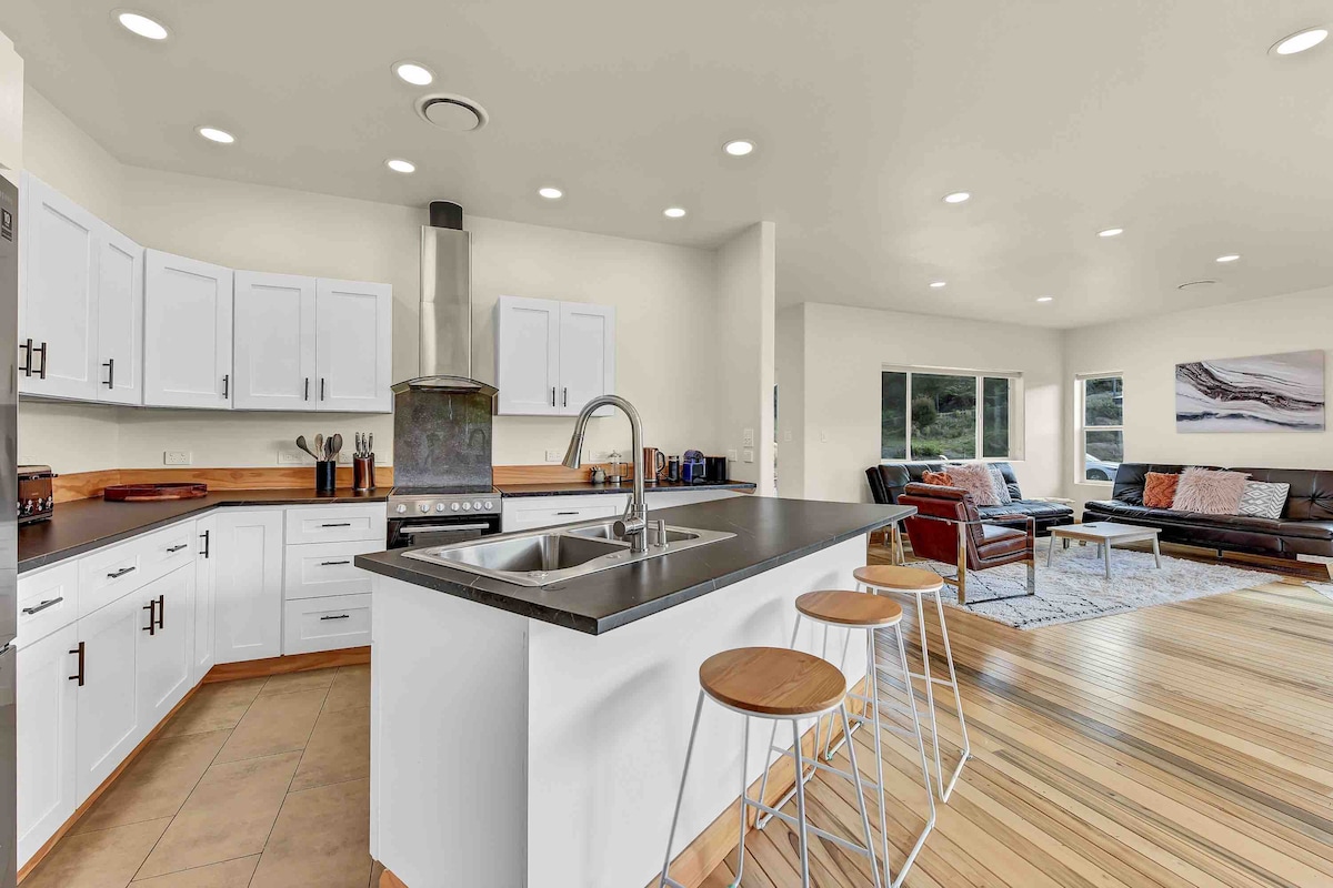 An open plan kitchen features modern white cabinetry paired with a sleek dark countertop. Three wooden stools are positioned at the edge of the island. The adjacent living area displays a comfortable seating arrangement with a muted color palette and large windows allowing natural light to fill the space.