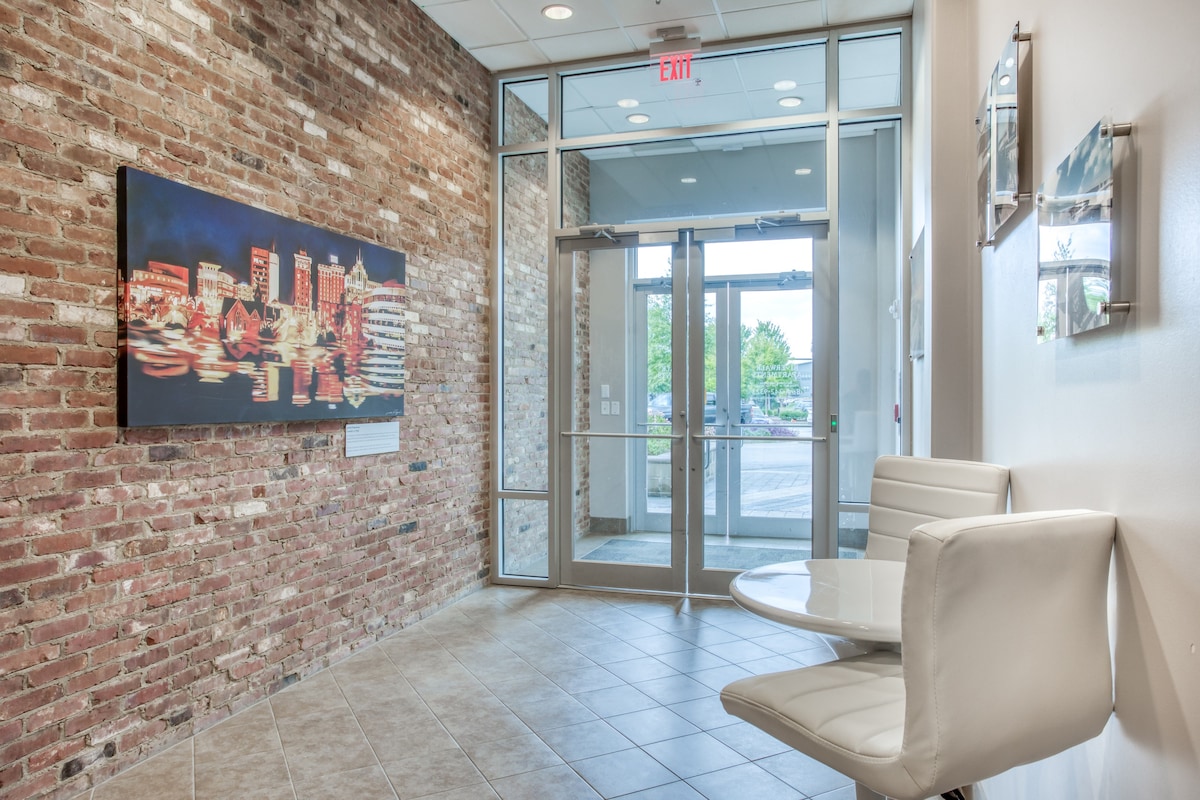 A well-lit entryway showcases a modern design with exposed brick walls. A contemporary artwork piece is displayed prominently on one wall. A round table with two white chairs is positioned in the corner, offering a welcoming space that leads to the glass doors opening to the outside.
