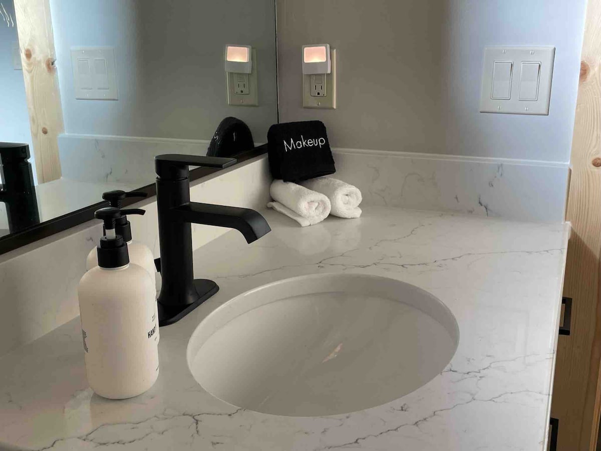 A modern bathroom sink area features a sleek black faucet and a circular basin set into marble countertop. Neatly arranged beside the sink are two fluffy white towels and a black makeup bag, complementing the sophisticated decor.