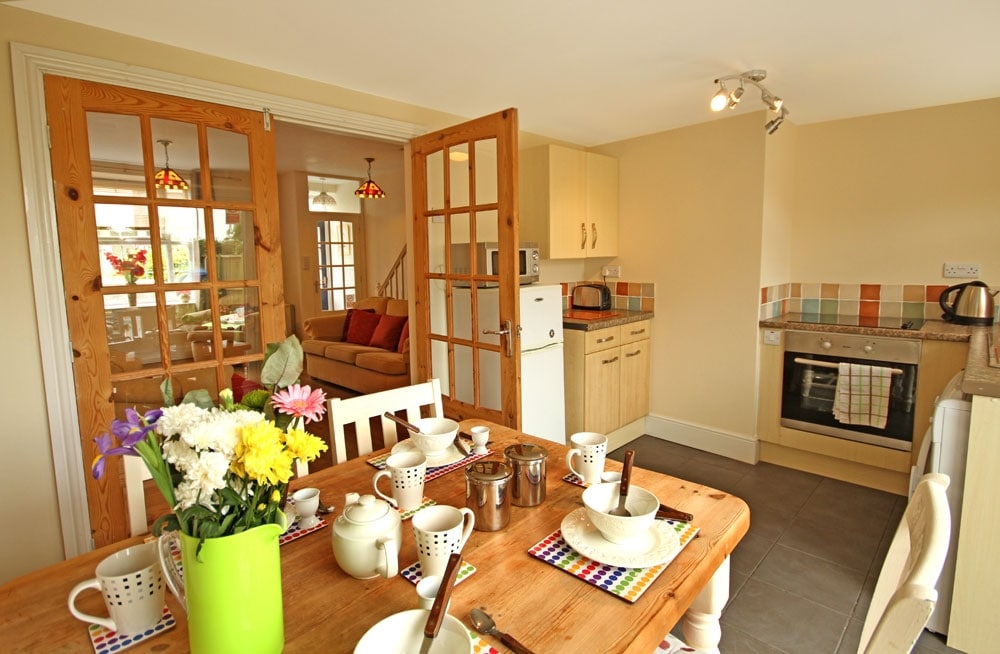 A bright kitchen area is visible, featuring a wooden dining table set with plates and utensils. Glass double doors lead to a living area. Light-colored cabinetry and colorful tiled backsplash complement the space. White flowers are arranged in a green vase on the table.