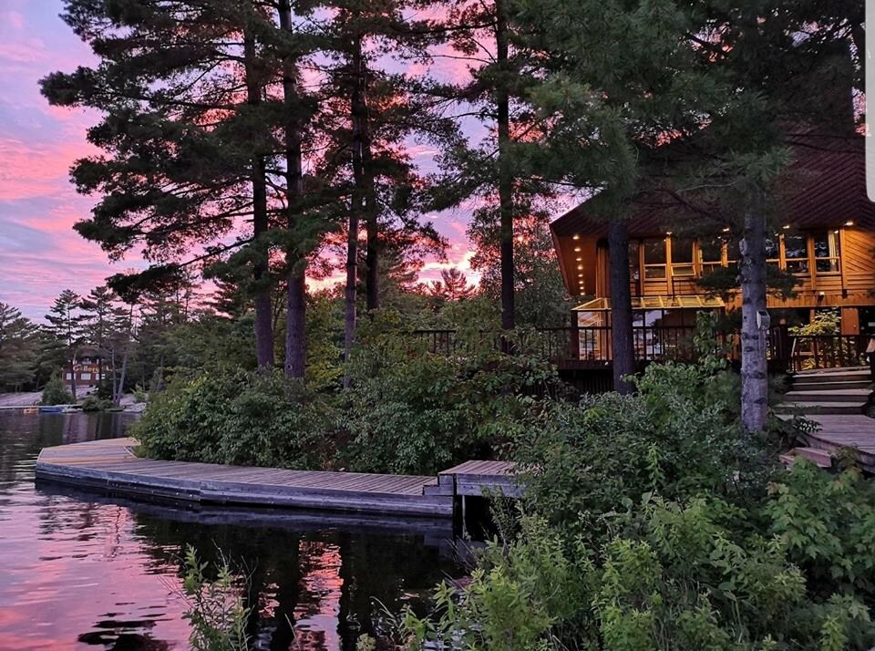 A scenic view captures a wooden deck extending over calm water, surrounded by lush greenery and tall trees. A cozy cabin is visible, featuring large windows illuminated by the warm glow of sunset. Soft pink and purple hues fill the sky.