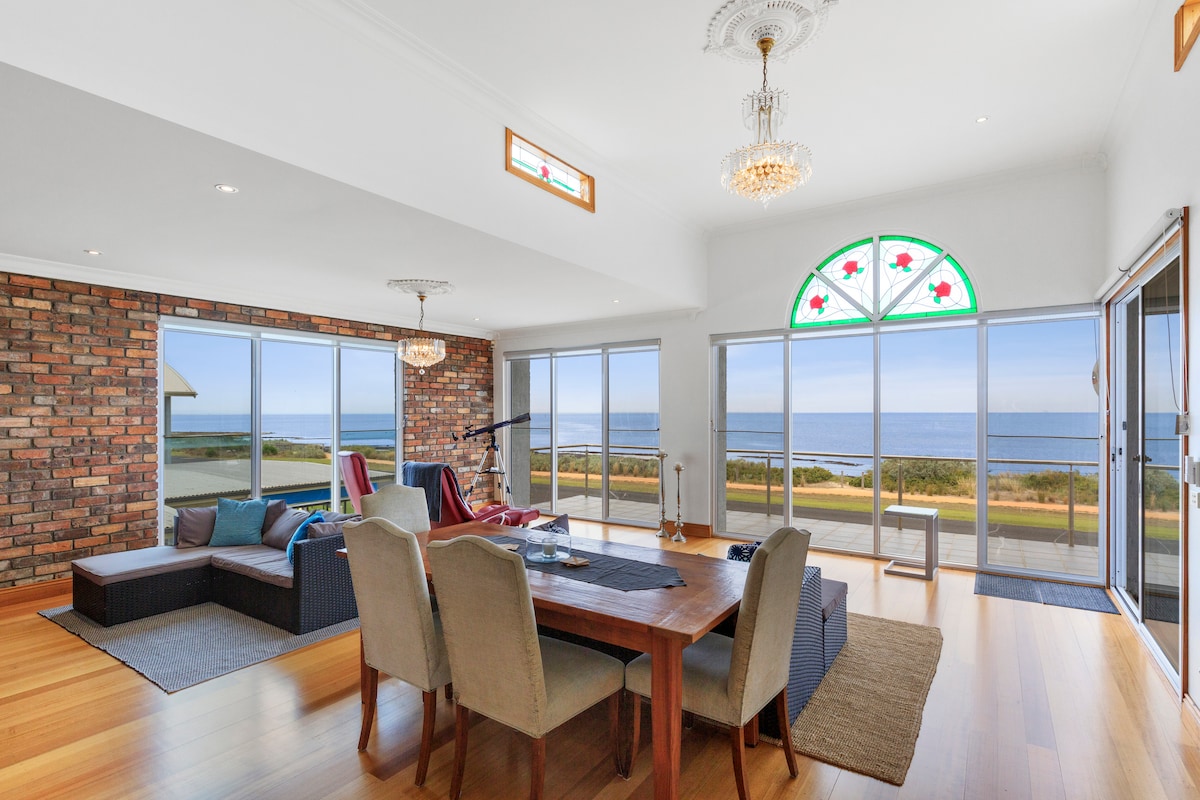 An open-plan living area features a long wooden dining table surrounded by upholstered chairs. Large windows offer panoramic views of the coastline, with natural light flooding the space. A cozy sectional sofa and a telescope invite relaxation while enjoying the serene seaside backdrop.
