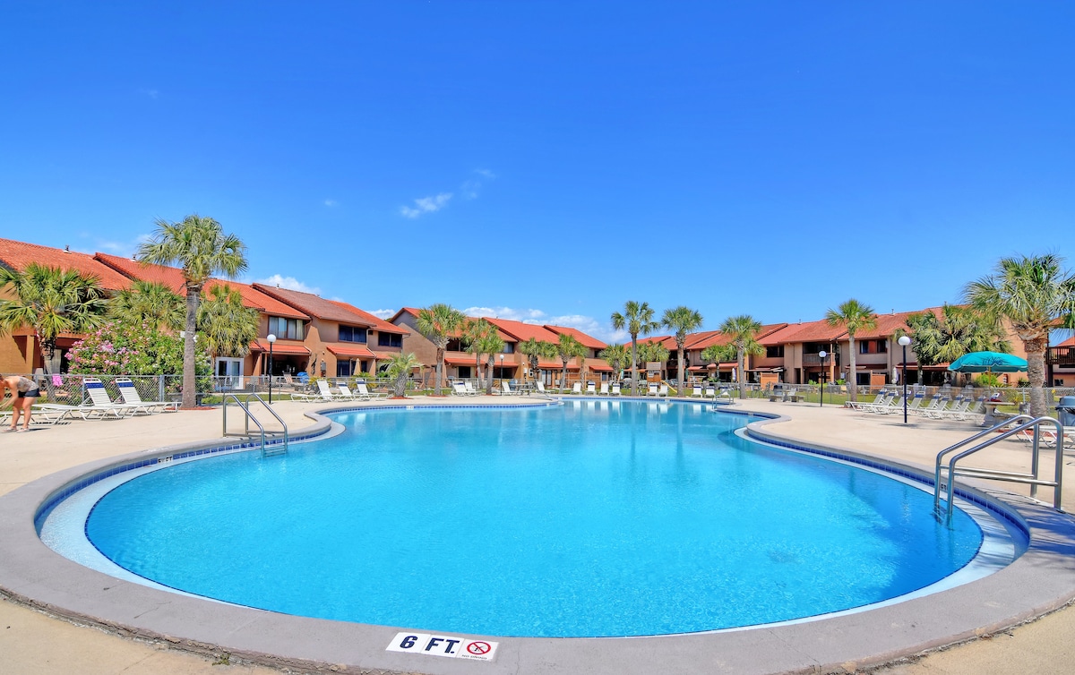 A large swimming pool is surrounded by lounge chairs under clear blue skies and palm trees. The pool features gentle curves and a depth marking of 6 feet. The backdrop includes colorful two-story buildings that enhance the resort atmosphere.