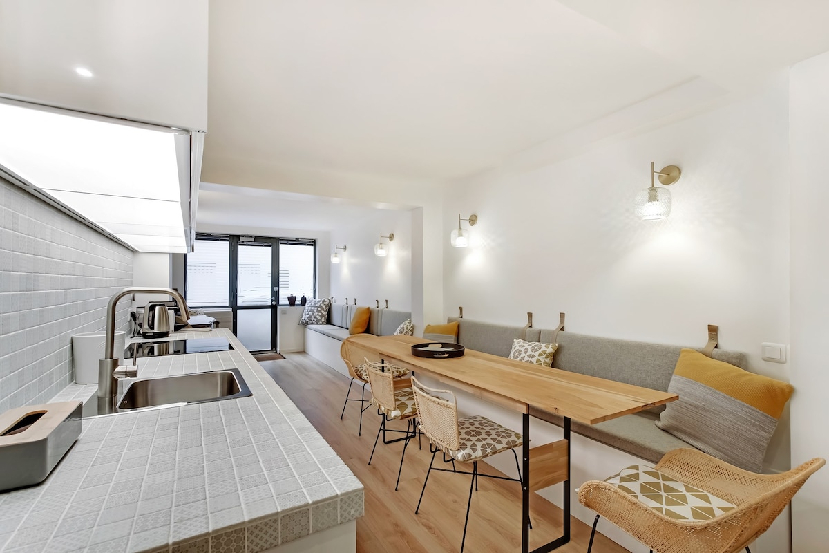 A spacious kitchen area features a wooden dining table adjacent to a cozy seating nook, both illuminated by soft lighting. A countertop with appliances is visible, complemented by a light-colored backsplash. Large windows allow natural light, with a view of a private outdoor space.