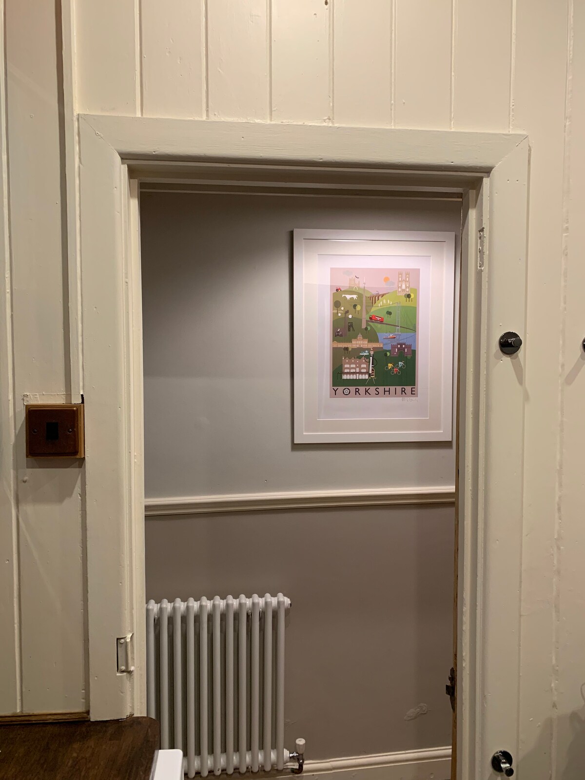 An interior doorway leads to a softly lit space featuring a framed artwork representing Yorkshire on the wall. A radiator is positioned beneath the artwork, while the wall displays a neutral color palette with a lower panel in a contrasting shade.