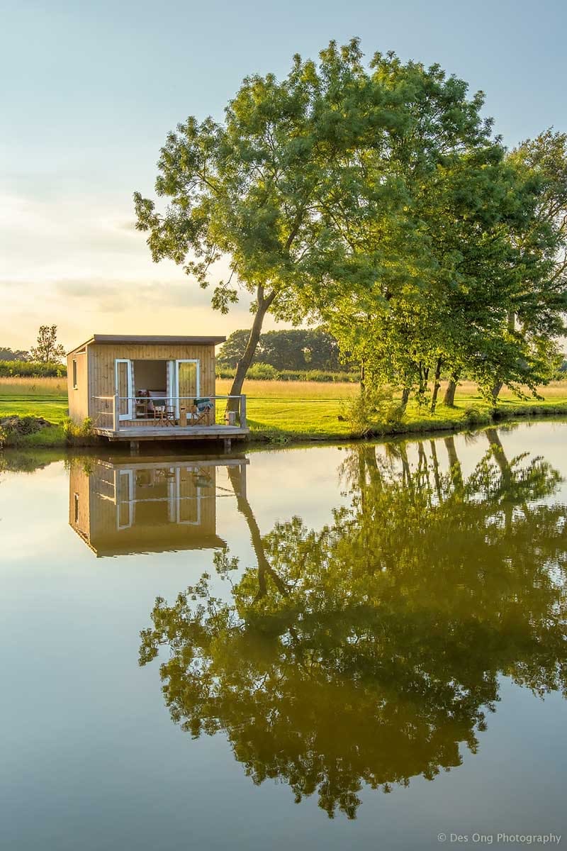 A cozy lakeside cabin is positioned at the water's edge, reflecting beautifully in the calm lake. Surrounding trees add a touch of nature, enhancing the peaceful outdoor setting. A small deck with seating invites relaxation while enjoying the tranquil views.