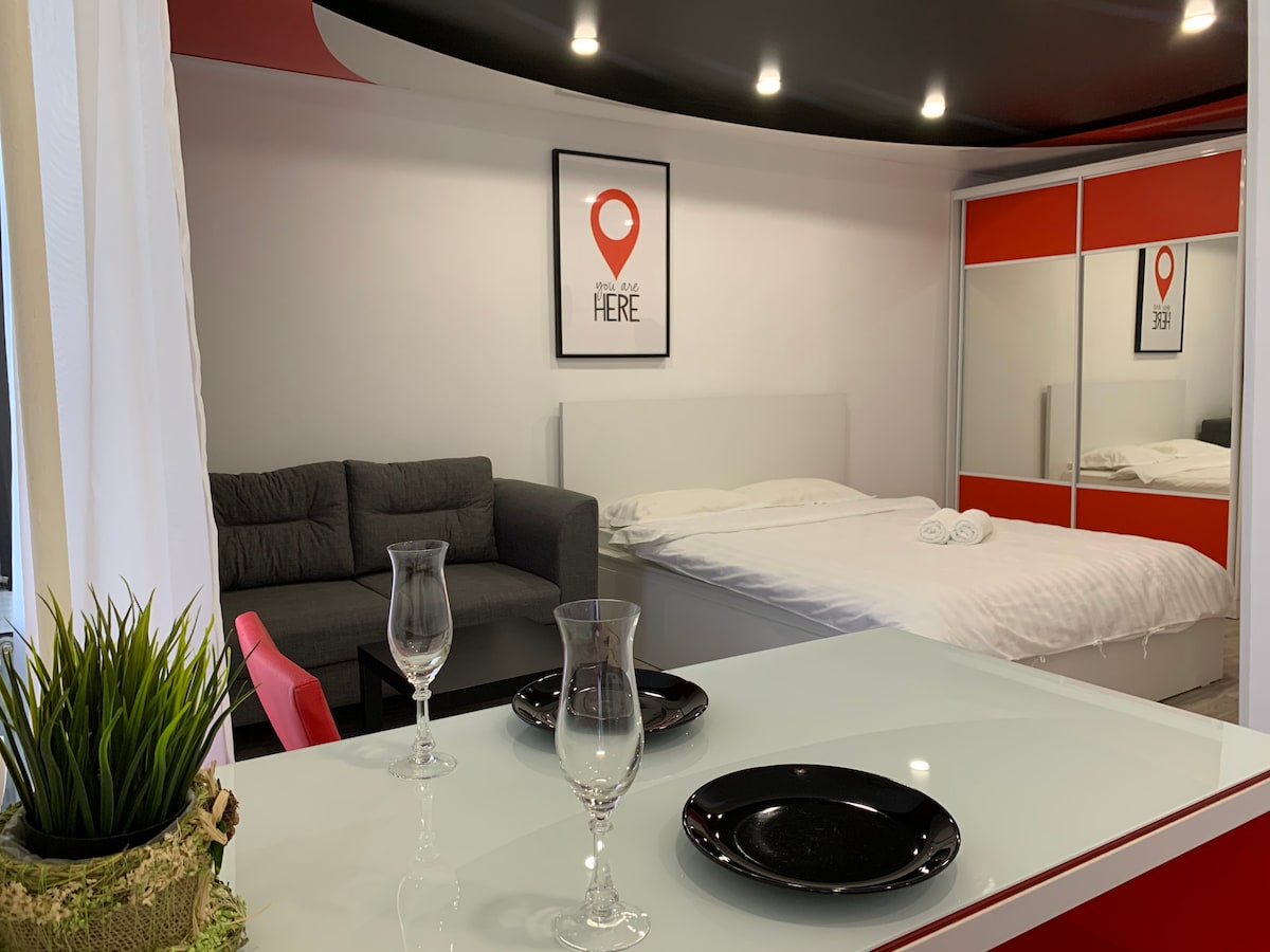 A studio apartment showcases a modern design with a cozy sleeping area featuring a spacious bed and a comfortable gray sofa. A dining table is set with black plates and glassware, while a large mirrored wardrobe reflects the stylish decor.