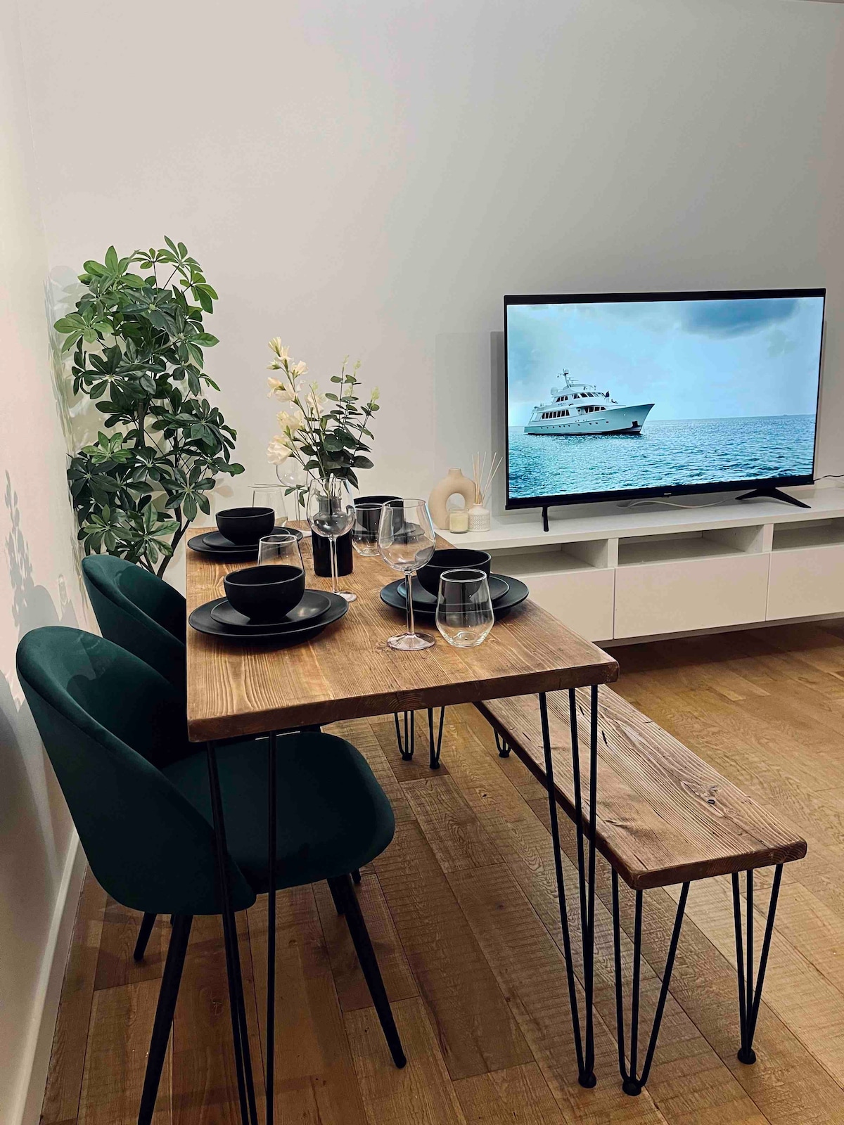 A dining area features a wooden table with four plates and glassware arranged neatly. Two green chairs accompany the table, while a television displays a serene marine scene. A potted plant adds a touch of greenery to the space.
