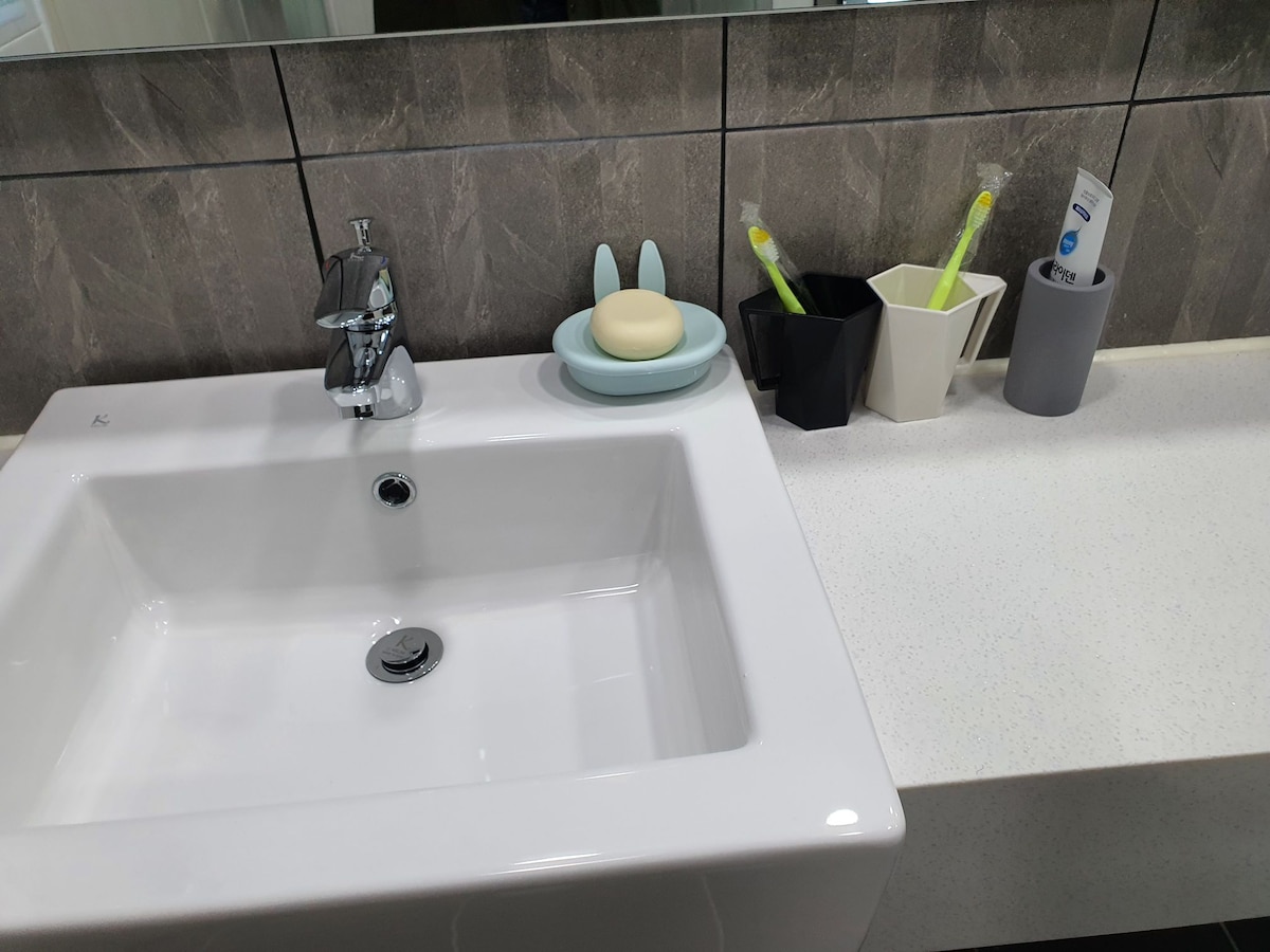 A clean bathroom sink featuring a glossy white basin and a modern faucet. Adjacent to it, a soap dish holds a bar of soap shaped like a bunny. Simple containers are arranged on the counter, holding toothbrushes and toiletries against a backdrop of textured gray tiles.