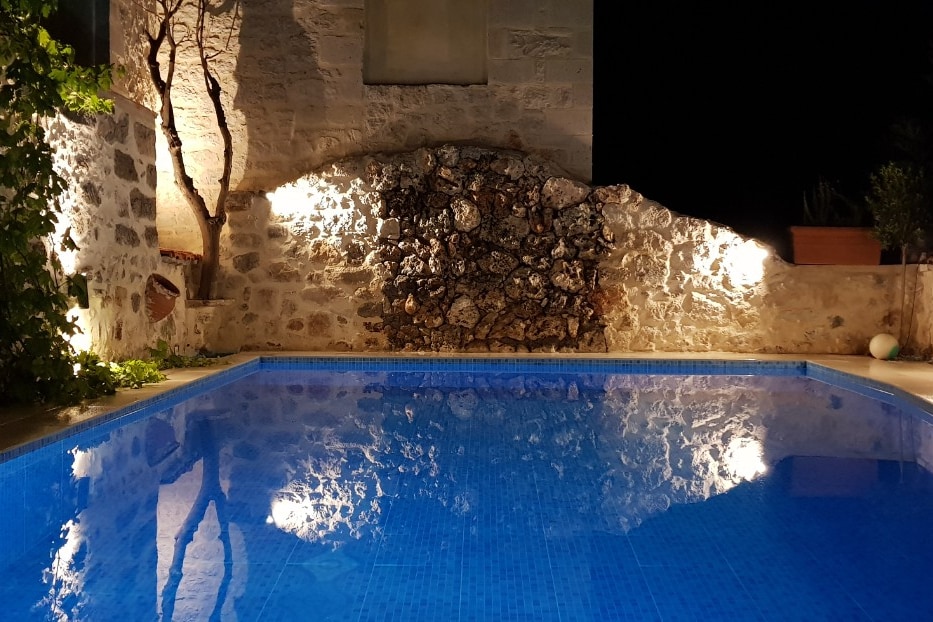 A private pool is illuminated under soft lights, surrounded by a stone wall and greenery. The tranquil water reflects the surrounding features, creating a serene atmosphere. The rustic charm of the stone structure is highlighted by the warm glow from the lights.
