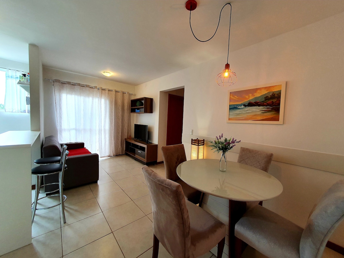 An open living space features a dining table surrounded by four upholstered chairs. A sofa is positioned nearby, with a television on a low cabinet across from it. Natural light is filtered through sheer curtains on a large window.
