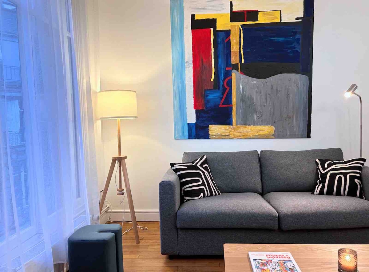 A modern living space is highlighted by a large, abstract painting on the wall. A gray sofa features patterned cushions, and is positioned near a wooden coffee table. Soft lighting is provided by a floor lamp and a table lamp, creating a cozy ambiance.