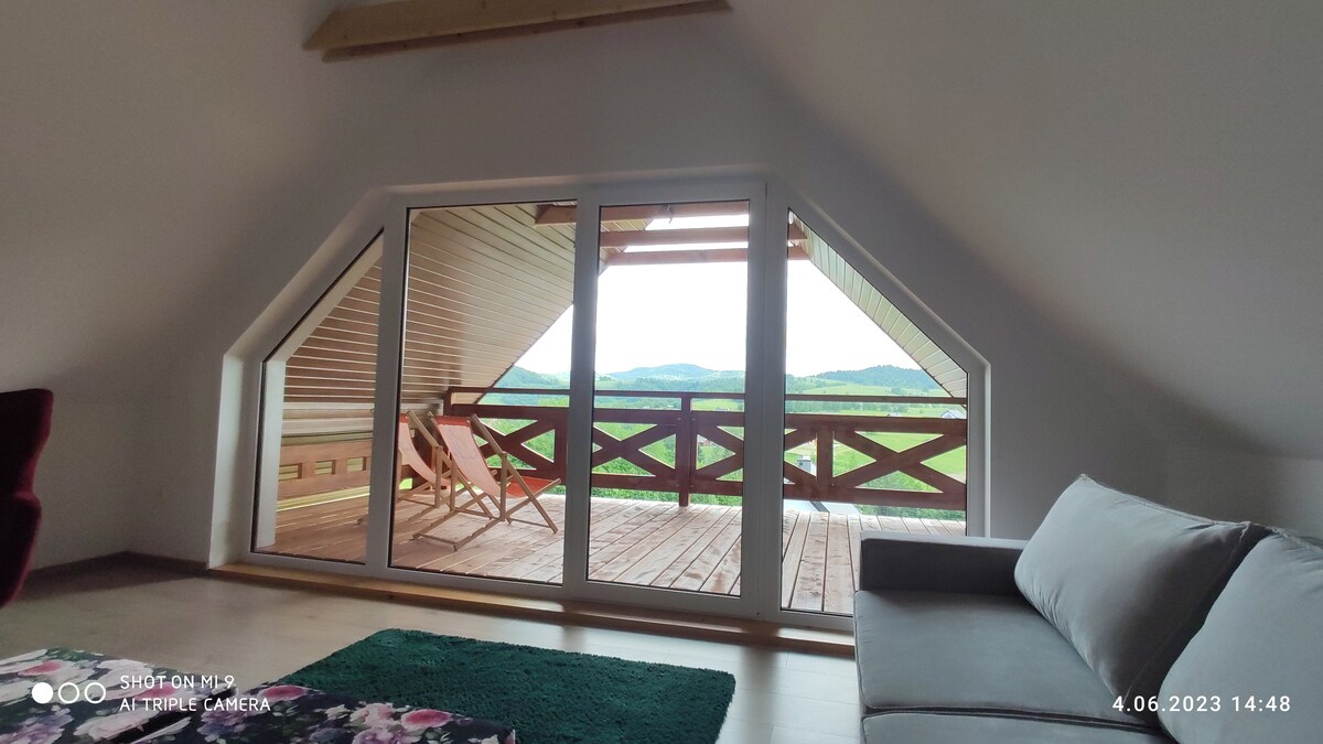 A spacious living area features a large window with panoramic views of the surrounding mountains. Sliding glass doors lead to a private balcony, where two lounge chairs invite relaxation. Natural light fills the room, highlighting the softly colored walls and cozy furnishings.