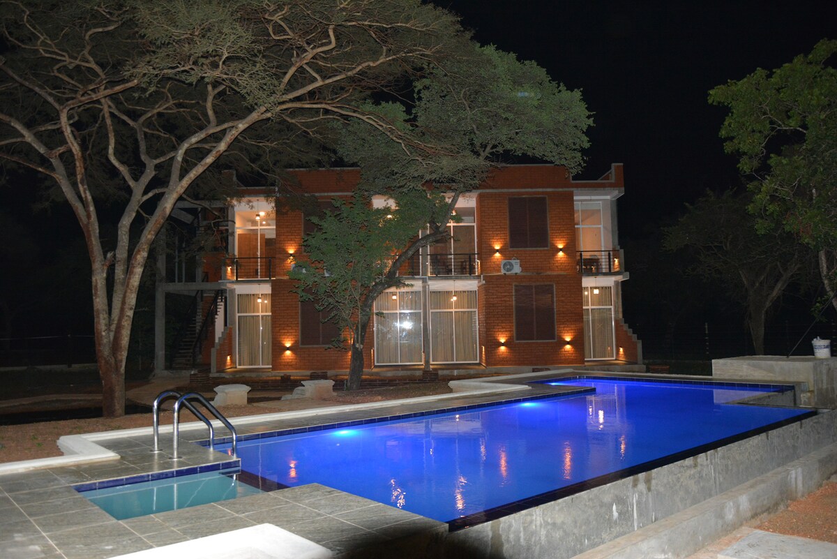 A modern two-story building is seen at night, featuring soft lighting that highlights its brick exterior. A spacious swimming pool with a refreshing blue hue reflects the surrounding trees and architecture, inviting relaxation under the night sky.