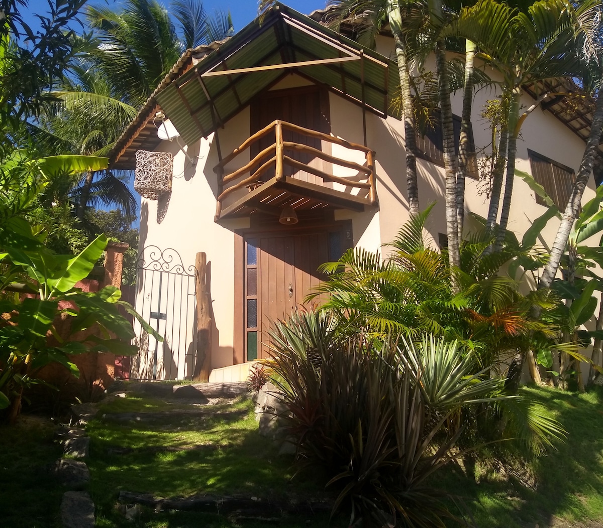 A charming chalet stands amidst lush greenery, featuring a wooden entrance door and a balcony with a rustic railing above. Tropical plants frame the entrance, enhancing the serene ambiance of the outdoor area.