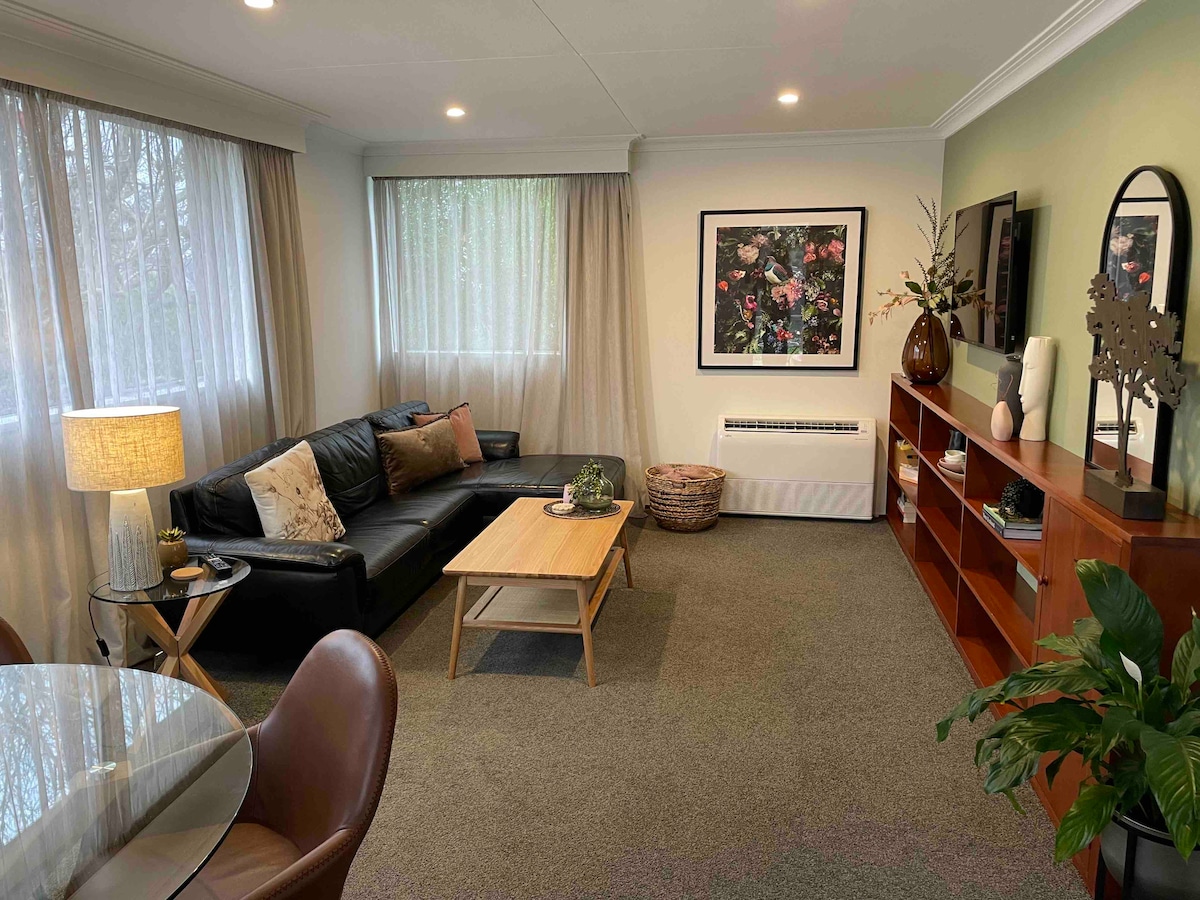 A stylish living area is presented with a black leather sofa, complemented by decorative cushions. A wooden coffee table sits at the center, while a large artwork adorns the wall, and natural light filters through sheer curtains, enhancing the relaxed ambiance.