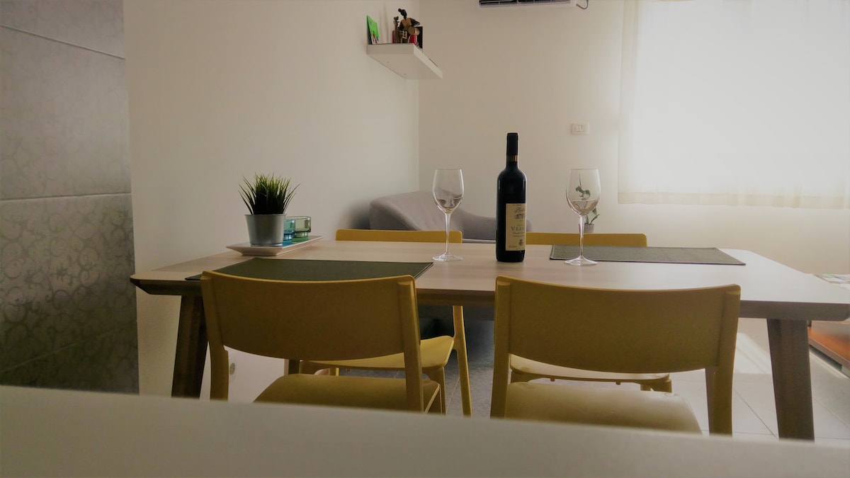 A dining area is shown with a light wooden table set for two. Two yellow chairs flank the table, which features a bottle of wine and two glasses. A soft gray sofa is visible in the background, with natural light illuminating the space through a nearby window.