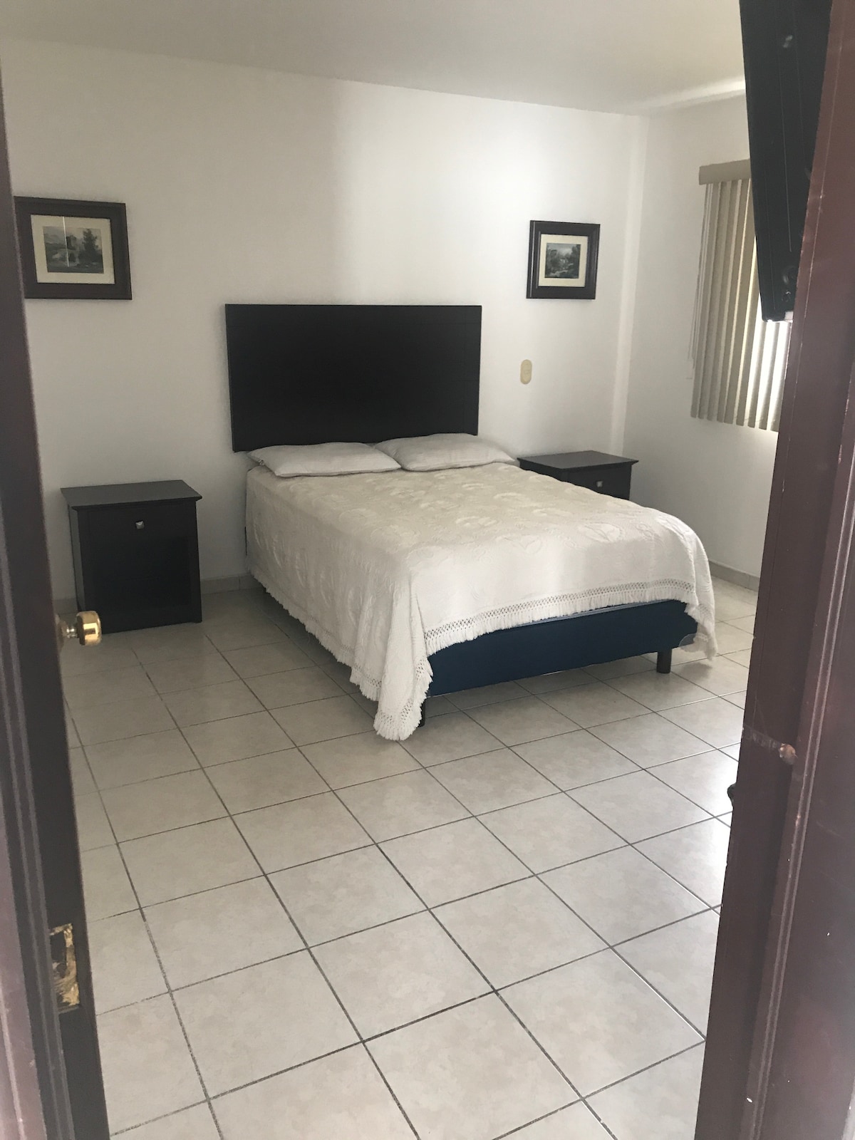 A private room is showcased, featuring a spacious bed with a white blanket. Two nightstands flank the bed, with framed artwork adorning the walls. Natural light enters through a window covered by vertical blinds, illuminating the tiled floor in a neutral tone.