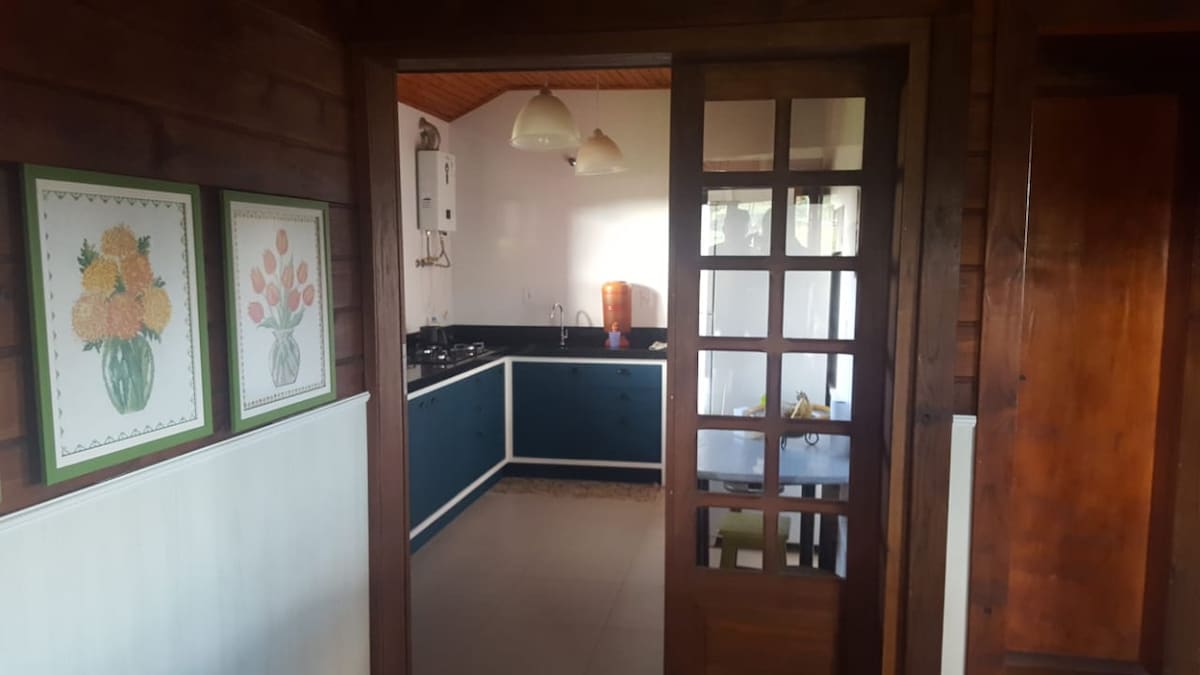 A modern kitchen is visible through a wooden door. The space features dark cabinets and a light countertop. Two pendant lights hang above the area. Floral artwork is displayed on the wall, adding a touch of colour to the room.