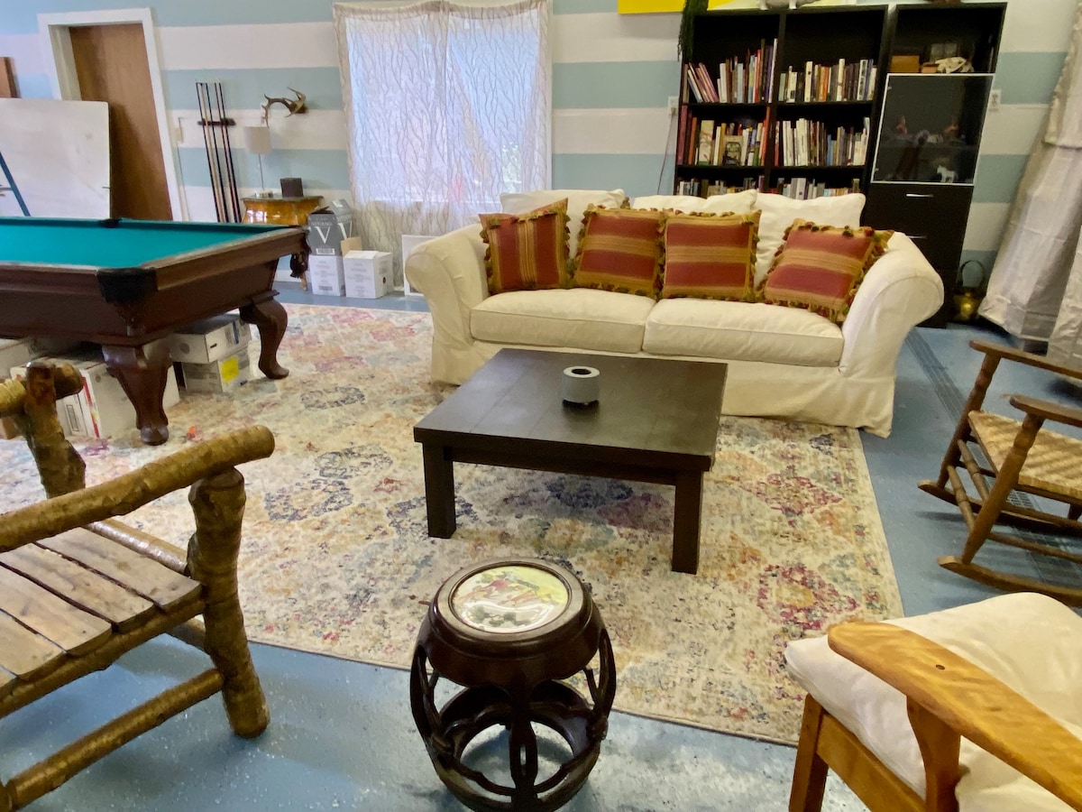 A spacious lounge area is presented with a large, cream-colored sofa adorned with decorative pillows. A dark wooden coffee table sits in front of it, complemented by a patterned area rug. A bookshelf filled with books is visible in the background, alongside a pool table.