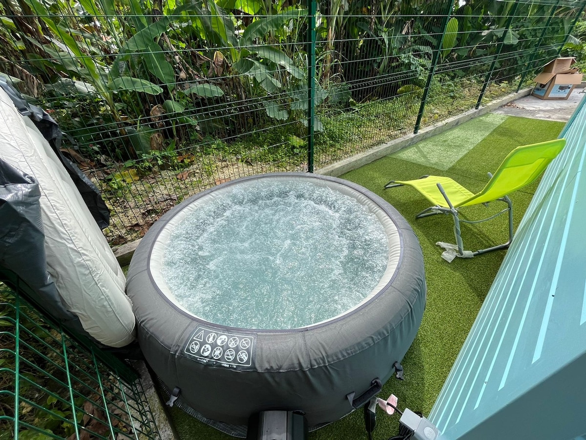 An inflatable jacuzzi is positioned on a green lawn-like surface, surrounded by lush greenery. A light green chair is situated nearby, offering a relaxing spot to unwind. The bubbling water of the jacuzzi invites relaxation after outdoor activities.