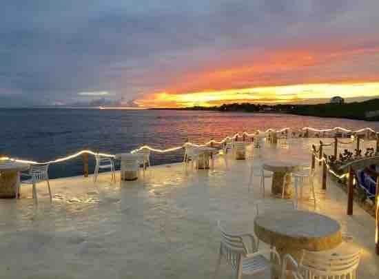 A serene waterfront view is captured during sunset, showcasing vibrant shades of orange and pink in the sky. A spacious patio is lined with round tables and white chairs, illuminated by soft string lights, creating a calm atmosphere by the water.