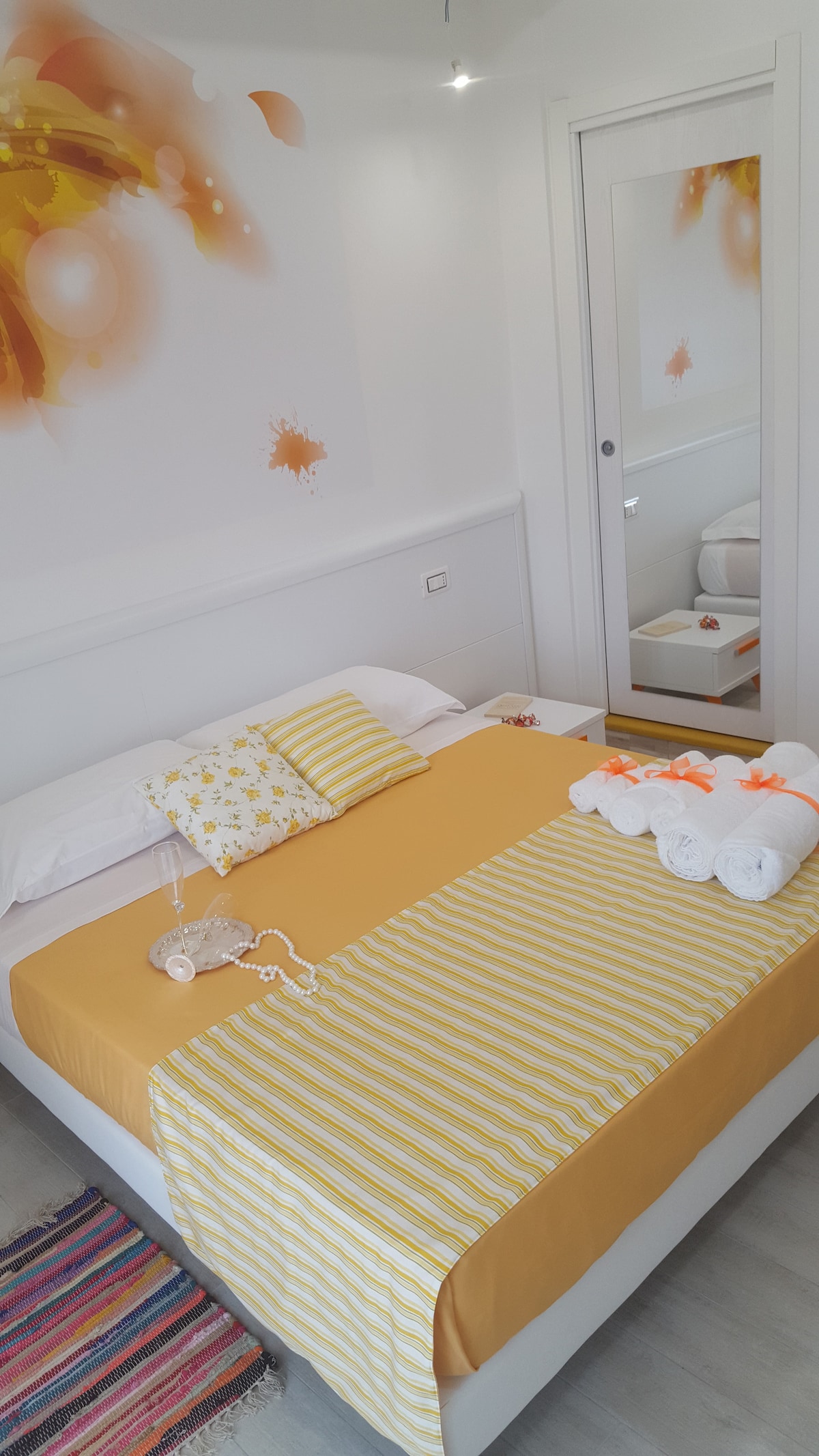 A bright sleeping area features a spacious bed with a vibrant yellow and white striped blanket, complemented by decorative cushions. Fresh towels are neatly arranged at the foot of the bed. A mirror reflects the room, and a small accessory table is positioned nearby.