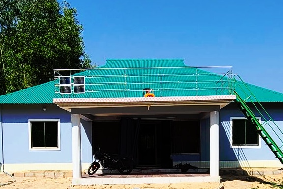A spacious building features a vibrant green roof and is complemented by a light blue exterior. Large windows allow natural light to enter. A rooftop area is accessible via a green staircase, with solar panels installed on the roof. A motorcycle is parked beneath the covered porch.