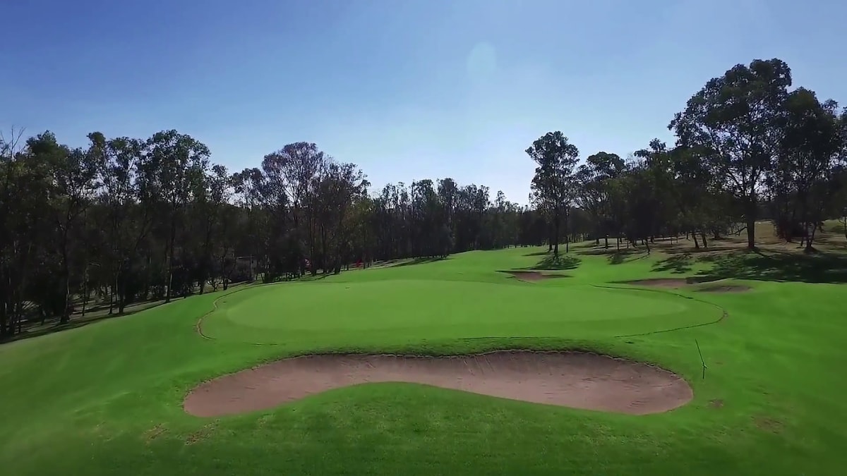 A well-maintained golf course is displayed, with a large green area surrounded by a smooth sand trap. Lush trees outline the perimeter, providing a natural backdrop against the clear blue sky. The layout emphasizes open spaces and well-groomed grass.