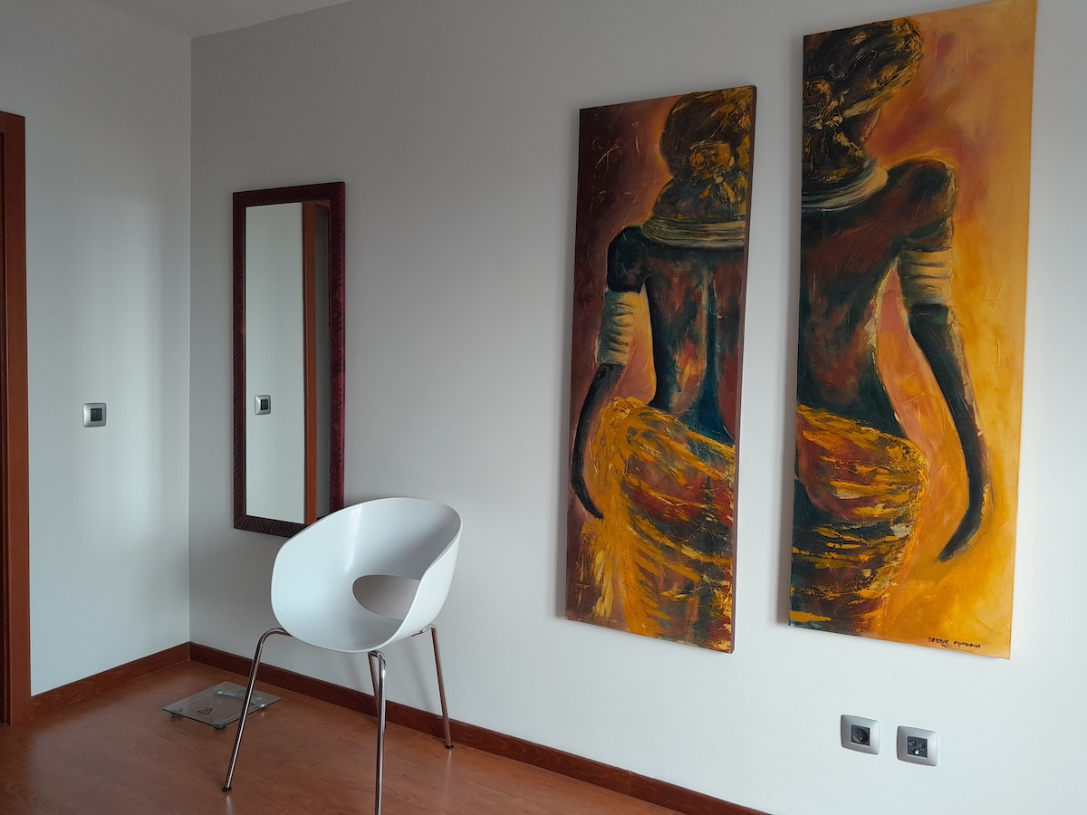 A corner of the apartment features two large paintings depicting figures in vibrant hues. A sleek white chair is positioned near a full-length mirror on the wall, and warm wooden flooring provides a contrasting element to the light-colored walls.